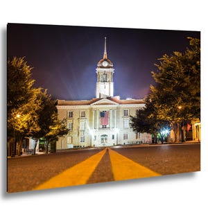 May include: A nighttime photograph of a historic building with a clock tower, lit by streetlights. The building has a large American flag hanging from the front. Yellow lines lead to the building.