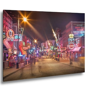 Pode incluir: Uma cena noturna de rua com letreiros de néon e figuras borradas. Os letreiros anunciam negócios como "Soul City Love" e "Beale Street". A imagem é uma representação vibrante da vida da cidade, com luzes brilhantes.