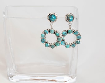 May include: Silver-tone earrings with turquoise stones. Each earring has a circular design with turquoise stones and a larger turquoise stone at the top. The earrings are set against a clear glass background.