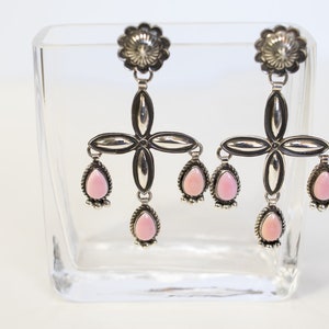 May include: A pair of silver earrings with pink stone accents. The earrings feature a cross design with dangling pink stones.