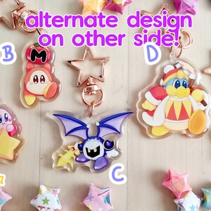 May include: Five different character keychains featuring characters from the video game Kirby. The keychains are made of clear acrylic with colorful printed images of the characters. The characters are Kirby, King Dedede, Meta Knight, Waddle Dee, and a pink Kirby holding a star. The keychains are hanging from gold-colored star-shaped clasps. The text "alternate design on other side!" is written in pink letters at the top of the image.