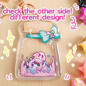 May include: A clear jar-shaped acrylic charm featuring a pink and blue cartoon squid inside. The jar has a light blue bow and a wooden lid. The text "check the other side! different design!" is written on the image. Surrounding the charm are origami stars.