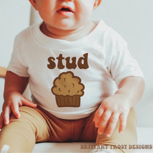 May include: A white baby onesie with brown text that says "stud" and a brown cartoon muffin illustration.