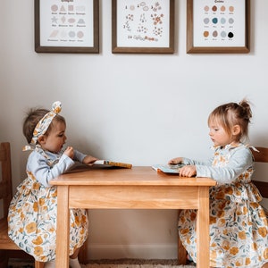 May include: Two young children sit at a small wooden table, each with a book. They wear matching floral dresses and headbands. Above them, three framed prints display shapes, numbers, and colors. The room has a neutral color palette with a patterned rug.