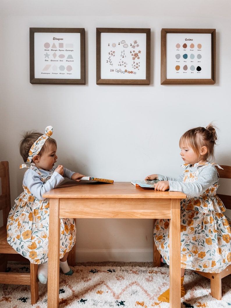 May include: Two young children sit at a small wooden table, each with a book. They wear matching floral dresses and headbands. Above them, three framed prints display shapes, numbers, and colors. The room has a neutral color palette with a patterned rug.