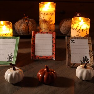 May include: Three rectangular notepads with lined paper, each featuring 'Happy Halloween' text and distinct designs: a green tree motif, an orange pumpkin pattern, and a brown spiderweb print. Three small pumpkins and three lit candles accompany them.