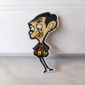 Könnte beinhalten: Gestickter Bügel-Patch mit einer Comicfigur in braunem Anzug, roter Krawatte und einem überraschten Gesichtsausdruck. Die Figur hat eine große Nase, schwarze Haare und weiße Augen.