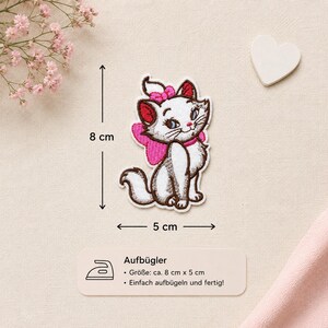 Könnte beinhalten: Ein gestickter Aufbügler in Form eines weißen Kätzchens mit blauen Augen, einer rosa Schleife auf dem Kopf und einer rosa Schleife am Rücken. Der Aufnäher misst 8 cm in der Höhe und 5 cm in der Breite, mit braunen Konturen und roten Ohrinnenseiten.