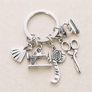 Könnte beinhalten: Ein silberfarbener Metallschlüsselanhänger mit fünf hängenden Anhängern: ein Kleid, eine Nähmaschine, ein aufgerolltes Maßband, eine Schere und ein kleines Bügeleisen. Die Anhänger sind an einem runden Schlüsselring aus Metall befestigt.