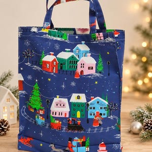Könnte beinhalten: Eine marineblaue Tragetasche mit einem festlichen Weihnachtsmotiv aus bunten Häusern, einem Weihnachtsbaum, einem Zug mit Geschenken und einer Pferdekutsche. Das Design zeigt weiße Schneeflocken und Sterne auf dunkelblauem Hintergrund.