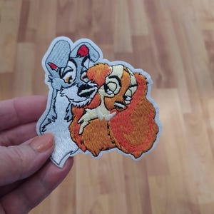 Könnte beinhalten: Gestickter Aufnäher mit Lady und der Tramp, einem grau-weißen Hund und einem braun-orangen Hund, die sich küssen. Der Aufnäher ist auf weißem Hintergrund.