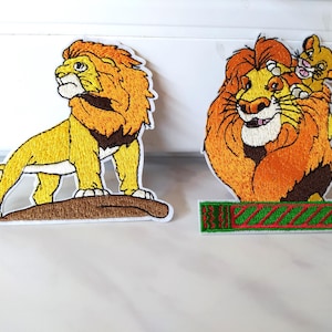 Könnte beinhalten: Zwei gestickte Bügelbilder mit den Figuren aus dem König der Löwen. Das erste Bügelbild zeigt Simba, der auf einem Felsen steht und nach oben schaut. Das zweite Bügelbild zeigt Mufasa und Simba zusammen. Beide Bügelbilder haben einen grün-roten Hintergrund.