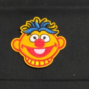 Könnte beinhalten: Ein gelber Filz-Stickerei-Aufnäher von Big Bird aus der Sesamstraße. Big Bird hat blaues und schwarzes Haar, eine rote Nase und ein breites Lächeln.