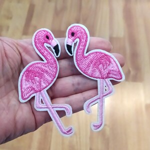 Könnte beinhalten: Zwei rosa Flamingo-Aufnäher zum Aufbügeln. Die Aufnäher sind mit rosa und weißem Garn gestickt.