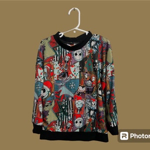 May include: A black and red long-sleeved sweater with a colorful print of characters from the Nightmare Before Christmas movie. The print includes Jack Skellington, Sally, and other characters from the film.