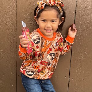 May include: A young person wearing a brown and orange long-sleeved shirt with a Halloween-themed print. The shirt features candy bars, pumpkins, and other Halloween imagery. The person is also wearing a brown headband with a bow.