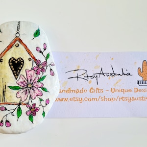 May include: Hand-painted, white stone featuring a birdhouse design with a heart-shaped opening, surrounded by pink flowers and green leaves. A small bird is perched above. A business card is next to the stone.