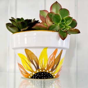 May include: White ceramic flower pot with a painted sunflower design in shades of yellow, orange, and brown. The pot is filled with green succulents.