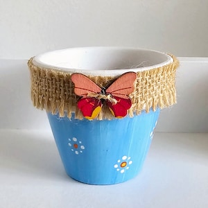 May include: A small blue planter with a white rim and a burlap trim. The planter has a painted design of white flowers with orange centres. A wooden butterfly with red and orange wings is attached to the burlap trim.