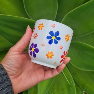 May include: A white ceramic cup featuring a hand-painted pattern of small, colourful flowers in shades of blue, purple, orange, red, and pink. The floral design is scattered across the exterior surface of the cup, which has a smooth, rounded shape.