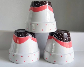Może przedstawiać: Trzy białe ceramiczne doniczki z wzorami w czarne i różowe kropki. Każda doniczka ma różową i czarną wstążkę wokół środka.