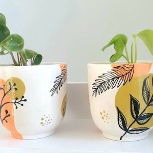 Könnte beinhalten: Zwei weiße Keramik-Blumentöpfe mit handgemalten abstrakten Mustern in Schwarz, Orange und Gold. Die Töpfe sind mit grünen Blattpflanzen gefüllt.