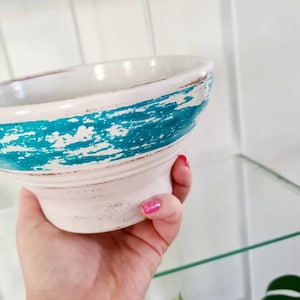 May include: A white ceramic planter with a turquoise blue band around the top. The planter has a distressed look with white paint showing through the turquoise.