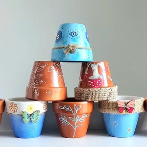 Puede incluir: Una colección de ocho macetas de terracota, cada una decorada con un diseño diferente. Las macetas están dispuestas en forma de pirámide, con la maceta más grande en la parte inferior y la más pequeña en la parte superior. Los diseños incluyen un ojo azul, hongos, hojas, mariposas y flores.
