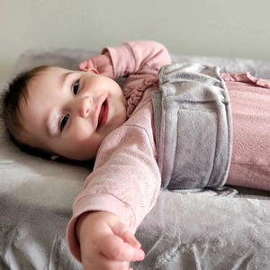 May include: A baby wearing a long-sleeved, light pink textured onesie with a ruffled neckline. A wide, soft, grey plush wrap is secured around the baby's midsection with a hook-and-loop fastener, resting on a matching grey plush surface.