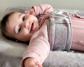 Könnte beinhalten: Ein Baby trägt einen langärmeligen, hellrosa strukturierten Body mit gerüschtem Ausschnitt. Ein breiter, weicher, grauer Plüschwickel ist mit einem Klettverschluss um die Körpermitte des Babys befestigt und liegt auf einer grauen Plüschunterlage.
