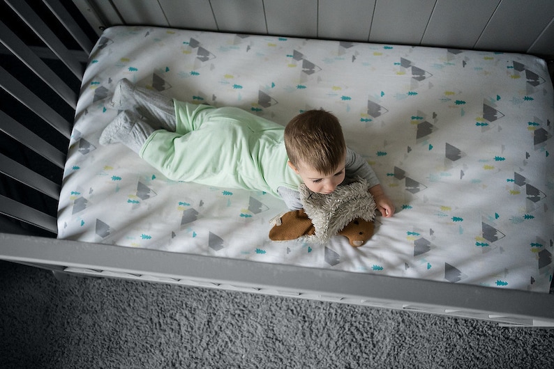 K&ouml;nnte beinhalten: Ein wei&szlig;es Bettlaken mit einem sich wiederholenden Muster aus grauen Bergen und kleinen t&uuml;rkisfarbenen sowie gelben Tannenb&auml;umen. Das Laken liegt auf einer Matratze in einem grauen Kinderbett, zusammen mit einem hellgr&uuml;nen Schlafsack und einem braunen Pl&uuml;schtier.
