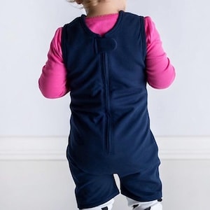 K&ouml;nnte beinhalten: Ein &auml;rmelloser dunkelblauer Strampler mit Rei&szlig;verschluss am R&uuml;cken, getragen &uuml;ber einem leuchtend pinken Langarmshirt und einer wei&szlig;en Leggings mit schwarzem Punktemuster. Der Strampler hat einen einfachen Rundhalsausschnitt und reicht bis zur Mitte der Oberschenkel.