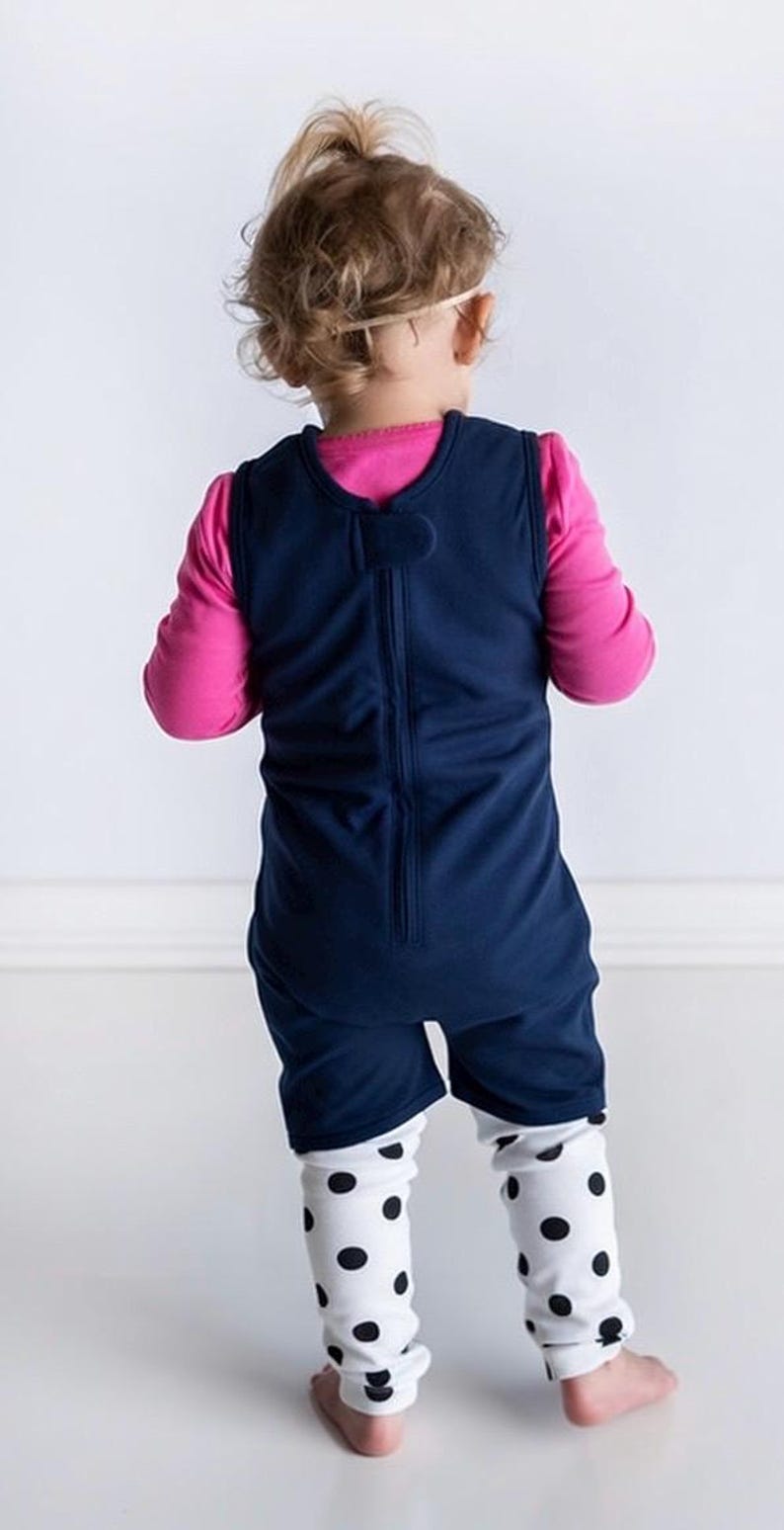 K&ouml;nnte beinhalten: Ein &auml;rmelloser dunkelblauer Strampler mit Rei&szlig;verschluss am R&uuml;cken, getragen &uuml;ber einem leuchtend pinken Langarmshirt und einer wei&szlig;en Leggings mit schwarzem Punktemuster. Der Strampler hat einen einfachen Rundhalsausschnitt und reicht bis zur Mitte der Oberschenkel.