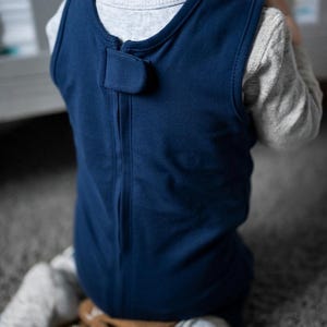 K&ouml;nnte beinhalten: Ein &auml;rmelloser, dunkelblauer Schlafsack mit einem Rei&szlig;verschluss auf der R&uuml;ckseite und einer kleinen Stofflasche, die am Halsausschnitt mit einem Klettverschluss gesichert ist. Das Kleidungsst&uuml;ck wird &uuml;ber einem hellgrauen Langarmshirt getragen.