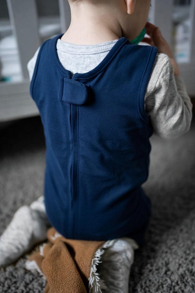 K&ouml;nnte beinhalten: Ein &auml;rmelloser, dunkelblauer Schlafsack mit einem Rei&szlig;verschluss auf der R&uuml;ckseite und einer kleinen Stofflasche, die am Halsausschnitt mit einem Klettverschluss gesichert ist. Das Kleidungsst&uuml;ck wird &uuml;ber einem hellgrauen Langarmshirt getragen.