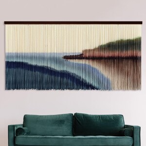 May include: A large rectangular wall hanging made of vertical cream-colored fiber strands. The fibers are dyed in a gradient pattern featuring shades of deep navy blue, light blue, and earthy brown, creating a landscape scene of a shoreline.