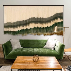 May include: A large wall hanging made of long, vertical cream-colored fiber strands. The lower sections of the strands are dyed in horizontal gradients of dark gray, forest green, and golden brown, creating a landscape-like pattern.