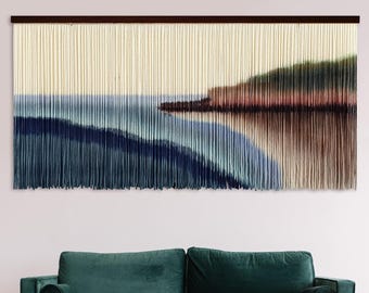 May include: A wall hanging with a woven tapestry design depicting a landscape with a body of water, a shoreline, and a hill in the background. The tapestry is made of yarn in shades of blue, green, brown, and beige.