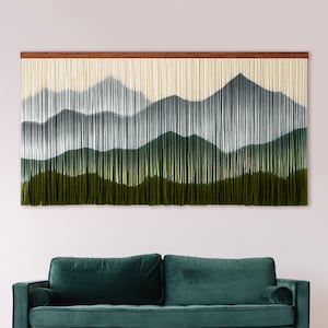 Könnte beinhalten: Ein großer rechteckiger Wandbehang mit einer Berglandschaft aus vertikalen Fasern. Das Design zeigt geschichtete Gipfel in den Farben Creme, Hellgrau, Dunkelgrau und tiefem Olivgrün, die an einer horizontalen Holzstange befestigt sind.