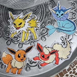 Può includere: Toppe ricamate raffiguranti quattro diversi personaggi Pokémon: Jolteon, Vaporeon, Eevee e Flareon. Le toppe sono colorate, con dettagli gialli, blu, arancioni e rossi. Sono posizionate su una superficie argentata decorativa.