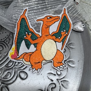 Op de afbeelding: Oranje en groene geborduurde strijk-op patch van een cartoon draak met vlammen op zijn staart.