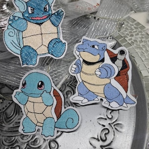 Peut inclure: Trois patchs brodés représentant différents stades de l'évolution du Pokémon Squirtle. Les patchs sont de couleur bleue, marron et crème et présentent la carapace emblématique du Pokémon.