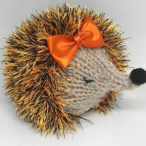 May include: A knitted hedgehog toy with a brown and orange spiky body and a beige face. The hedgehog has a large orange bow tied around its head and a black nose.