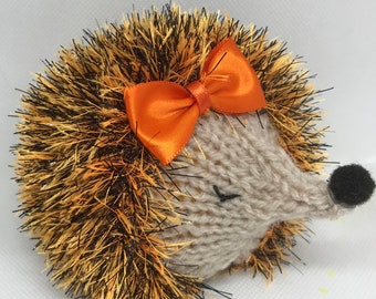 May include: A knitted hedgehog toy featuring a beige face with a black nose and closed eyes. The body is covered in textured, spiky yarn in shades of black and orange with a metallic sheen. An orange satin ribbon bow is attached to the top.