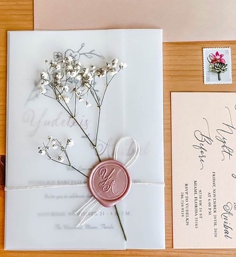 Puede incluir: Un conjunto de invitaciones de boda con una invitaci&oacute;n transl&uacute;cida con delicados detalles florales y un sello de cera rosa. La invitaci&oacute;n est&aacute; atada con una cinta blanca e incluye los nombres "Yudetris" y "Anibal". Tambi&eacute;n se ve una tarjeta a juego y un sello floral.