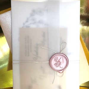 Puede incluir: Un conjunto de invitaciones de boda con una envoltura de vitela transl&uacute;cida, atada con hilo y sellada con un sello de cera rosa. El sello tiene las iniciales "Y" y "K" en una escritura elegante. La invitaci&oacute;n est&aacute; superpuesta a una tarjeta color crema.