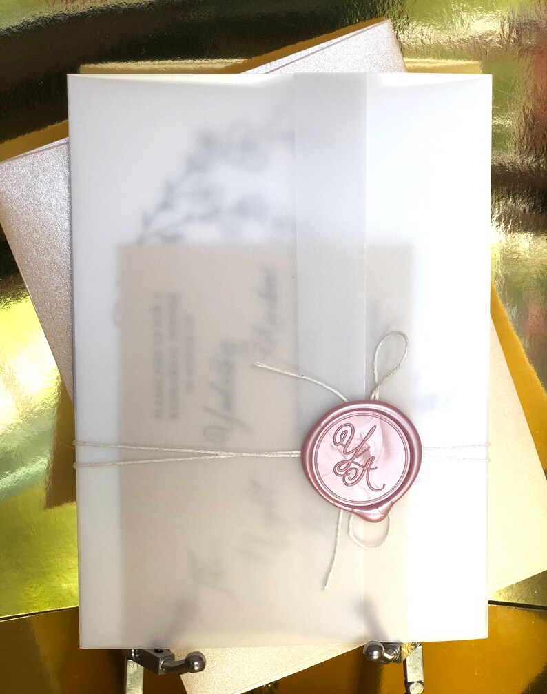 Puede incluir: Un conjunto de invitaciones de boda con una envoltura de vitela transl&uacute;cida, atada con hilo y sellada con un sello de cera rosa. El sello tiene las iniciales "Y" y "K" en una escritura elegante. La invitaci&oacute;n est&aacute; superpuesta a una tarjeta color crema.