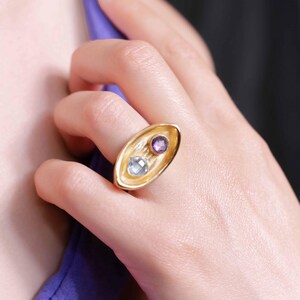 A woman displays a bold gold plated ring 'Gemship' with a square amethyst and round topaz, emphasizing its handcrafted beauty and creative style.