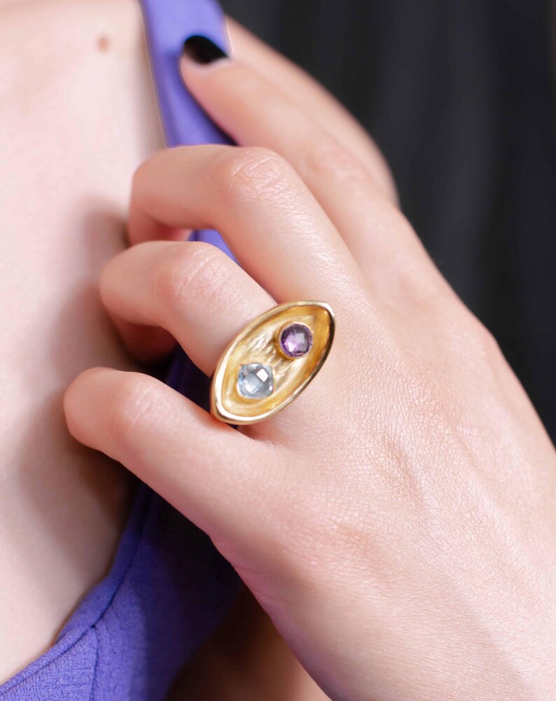 A woman displays a bold gold plated ring 'Gemship' with a square amethyst and round topaz, emphasizing its handcrafted beauty and creative style.