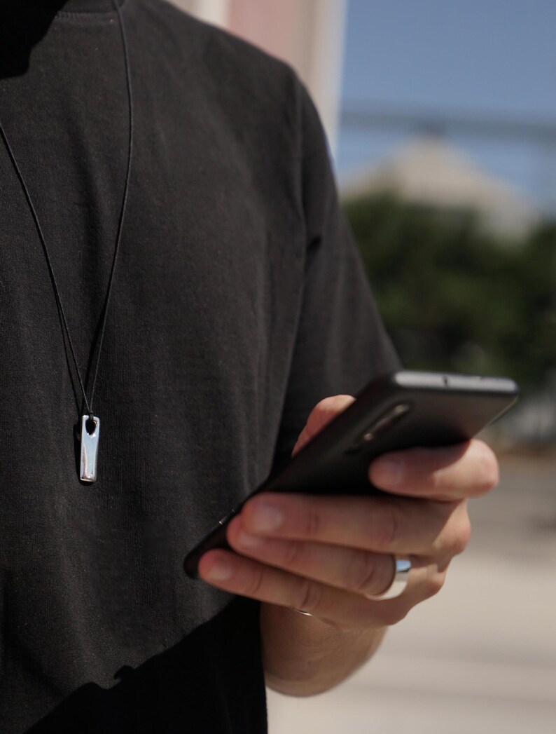 A man in a black shirt holds a cell phone, showcasing a handcrafted men's bar necklace in polished silver – perfect for modern style and everyday wear.